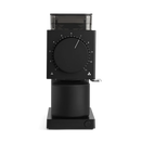 A sleek black manual coffee grinder with a large central dial, transparent bean hopper, and metal crank handle, shown on a clean white background.